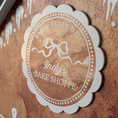 Logo of Indy's Bake Shoppe on a brown background with white decorative elements.